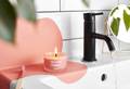 The candle is shown on a sink next to a running tap, that's partly coloured the same pinky orange as the candle's wax.