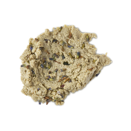 A close-up, top-down studio shot of a light tan, crumbly, dough-like facial cleanser (Lush's Angels on Bare Skin) spread in a dollop on a neutral background. The textured paste is visibly studded with small, dried purple and brown lavender buds