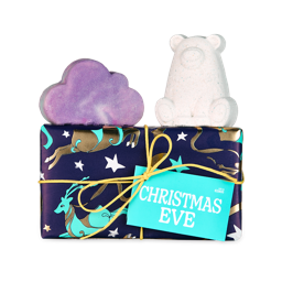 Christmas Eve. A short, rectangular gift box wrapped in dark blue paper with shiny, gold reindeer and tied in a yellow string with a teal gift tag. There is a cloud-shaped, purple bubble bar and a white polar bear-shaped bath bomb sitting on top.