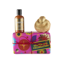 Serpentine Celebration. A rectangular gift box wrapped in colourful pink paper with stars and tied with a red string and gift tag. A bottle with golden shower gel and gold label sits on top of the box with a golden, coiled, snake-shaped bath bomb.